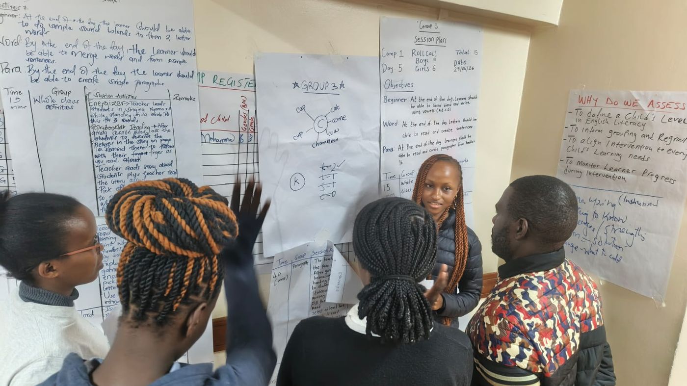 Team members discussing a poster titled 'Why Do We Assess' that sets out the M&E rationale